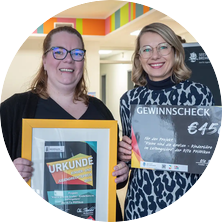 Das Foto zeigt zwei Frauen. Links Virginia Mibert. Rechts Cassandra Fee Berndt. Sie halten einen Gewinnscheck und eine Urkunde in ihren Händen.
